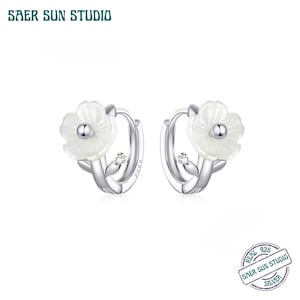 May include: A pair of silver hoop earrings with a flower design. Each earring features a white flower with a small silver bead in the center and a small clear gem. The earrings are from SAER SUN STUDIO.