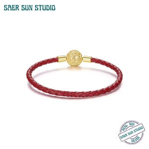 Puede incluir: Una pulsera de cuero trenzado rojo con detalles dorados. La pulsera presenta un cierre dorado con un diseño grabado. El nombre de la marca "SAER SUN STUDIO" es visible en la esquina superior izquierda de la imagen.