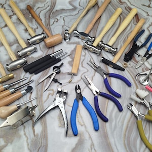 May include: An assortment of jewelry-making tools, including hammers, pliers, and other hand tools. The tools have wooden handles and metal heads, with various shapes and sizes. The tools are arranged on a marbled surface.