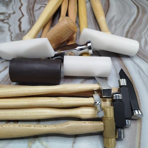 May include: A selection of woodworking tools, including hammers with wooden handles, white and black mallet heads, and metal components. The tools are arranged on a marbled surface.