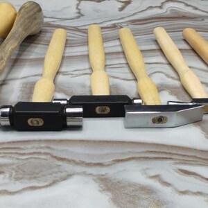 May include: A selection of metalworking tools, including hammers with wooden handles and various head shapes. There are also small, polished metal tools with rounded tops and flat bases. The tools are arranged on a light-coloured, marbled surface.