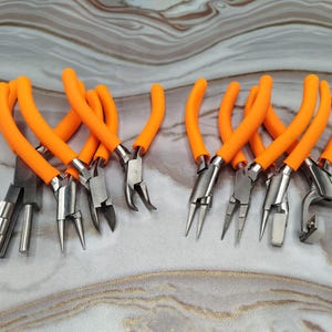Può includere: Una collezione di pinze per gioielli con manici arancioni brillanti. Le pinze sono realizzate in metallo argentato e includono vari stili come pinze a punta tonda, a punta piatta e a punta piegata. Gli strumenti sono disposti su una superficie a motivi marmorei.