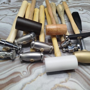 May include: A selection of hammers and tools, including wooden mallets, metal hammers, and a white cylindrical tool. The hammers have wooden and metal handles, with some featuring black or gold heads. The tools are arranged on a marbled surface.