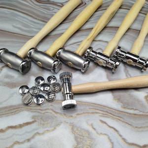 May include: An assortment of metal hammers with wooden handles and different textured heads, displayed with a selection of small, patterned metal discs. The tools are arranged on a marbled surface, suggesting a craft or jewellery-making theme.