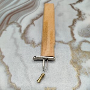 May include: A craft tool featuring a rectangular light-colored wooden handle attached to a silver-toned metal base. A thin, angled metal wire extends from the base, ending in a small, hollow, cylindrical brass-toned tip for precision work.