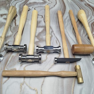 May include: A collection of various hammers with wooden handles and metal heads. The hammers have different head shapes, including ball-peen, flat, and rounded. The handles are light-colored wood, and the heads are silver or brass.