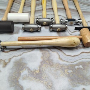May include: A selection of hammers with wooden handles and diverse head shapes. The collection features hammers with metal, rubber, and wooden heads. The hammers are displayed on a marbled surface, highlighting their varied designs and sizes.
