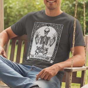 May include: Dark grey t-shirt with a white graphic of a skeleton electrician holding lightning bolts. The text "THE ELECTRICIAN" is below the graphic. The shirt is worn by a person.