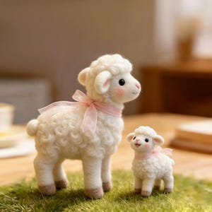 May include: Two needle-felted sheep figurines made of white wool with curly texture. The larger sheep features a light pink sheer ribbon tied around its neck, while the smaller sheep also wears a matching pink ribbon. Both have pink cheeks.