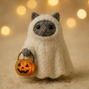 May include: A needle-felted gray cat figurine wearing a cream-colored ghost costume. The cat has dark eyes and a small pink nose. It is holding a miniature orange felted pumpkin bucket with a black jack-o'-lantern face carved into the front.
