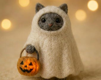 Pode incluir: Uma estatueta de gato cinzento em feltro, vestida com um traje de fantasma cor de creme. O gato tem olhos escuros e um pequeno nariz cor-de-rosa. Segura um balde de abóbora laranja em miniatura com uma cara de jack-o'-lantern preta.
