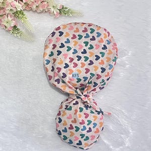 May include: A scrub cap with a colorful heart pattern in shades of pink, blue, green, and yellow. The cap is tied at the back and features a matching fabric design. The background is a soft, white, textured surface.