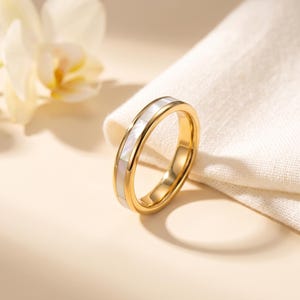 May include: A gold-toned band ring featuring a continuous inlay of rectangular white mother-of-pearl segments. The ring has a polished metallic finish and a smooth, rounded profile, showcasing a clean and simple design with a reflective surface.