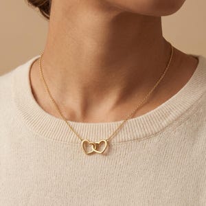 Puede incluir: Un collar dorado con dos colgantes entrelazados en forma de corazón. La fina cadena y los dijes de corazón destacan sobre un fondo neutro, resaltando el diseño de la joya. Un accesorio sencillo pero elegante.