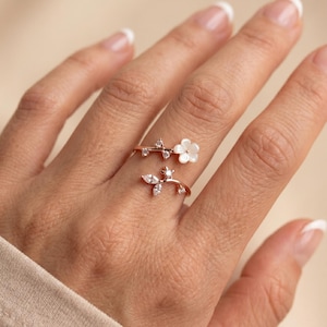 Puede incluir: Un anillo de oro rosa con un diseño floral. El anillo presenta una flor blanca y pequeñas piedras preciosas transparentes. El anillo se envuelve alrededor del dedo con una estética delicada inspirada en la naturaleza.