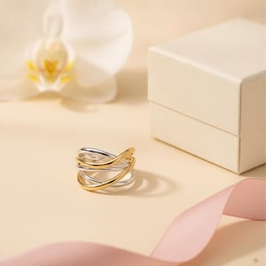 Puede incluir: Un anillo de varias bandas que presenta una combinación de hilos metálicos pulidos en tonos plateados y dorados que se entrelazan y superponen para crear un diseño continuo y en capas. El anillo tiene un acabado liso y una estructura abierta.