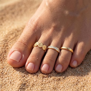 May include: Three gold-toned toe rings worn on three toes. The first ring features a series of small, connected flower shapes. The second ring is a band set with a row of small, clear, sparkling stones. The third ring is a plain, smooth gold band.