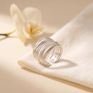 May include: A silver ring featuring a wide, multi-layered band design composed of several thin, overlapping, and intertwined metallic strands. The interior of the band is clearly stamped with the hallmark S925, indicating its sterling silver composition.