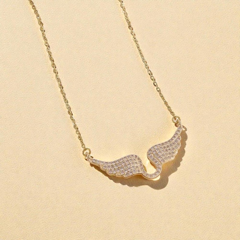 K&ouml;nnte beinhalten: Eine goldfarbene Halskette mit einem Anh&auml;nger in Form von Engelsfl&uuml;geln. Die Fl&uuml;gel sind mit kleinen, funkelnden Kristallen besetzt. Die Kette ist zart, und die Halskette ist vor einem hellbeigen Hintergrund platziert.
