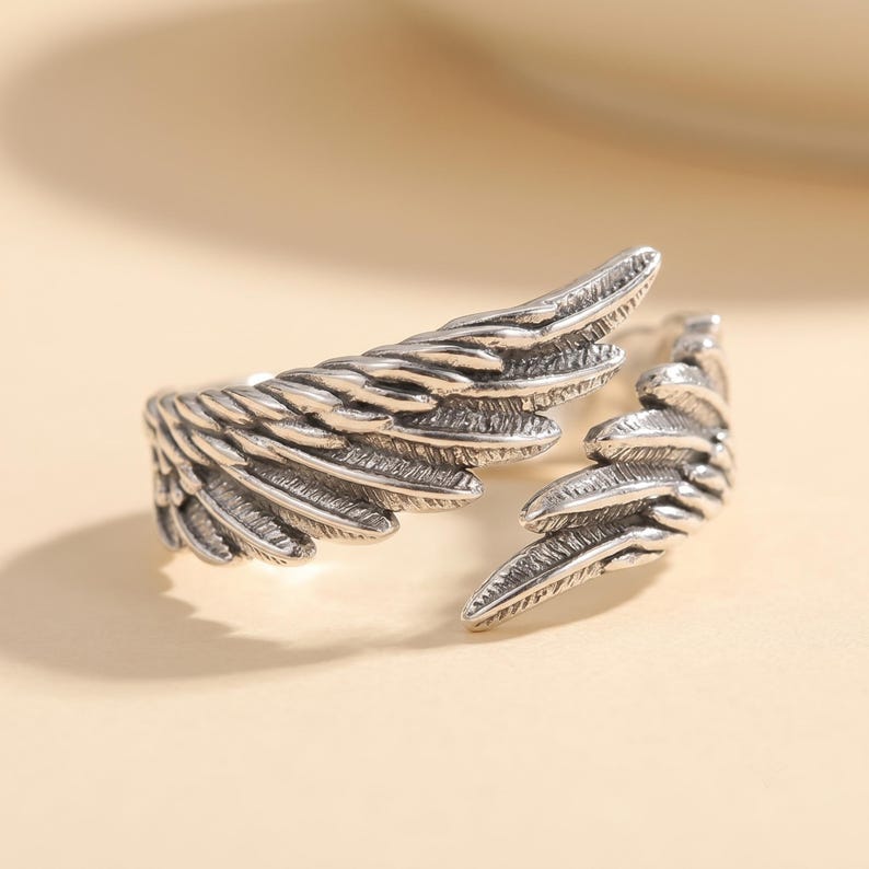 Pode incluir: Um anel aberto de tom prateado com dois designs detalhados de asas de anjo sobrepostas. O metal tem um acabamento oxidado que destaca os padr&otilde;es de penas texturizados na superf&iacute;cie das asas, criando uma apar&ecirc;ncia dimensional.