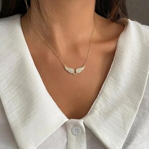 K&ouml;nnte beinhalten: Eine goldfarbene Halskette mit einem Anh&auml;nger in Form von Engelsfl&uuml;geln. Die Fl&uuml;gel sind mit kleinen, funkelnden Kristallen besetzt. Die Halskette wird vor einem wei&szlig;en Hemd mit Kragen gezeigt.