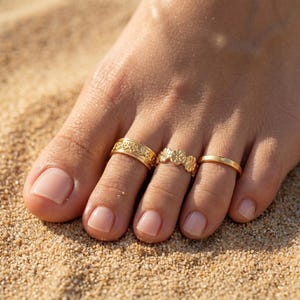 May include: Close-up of three gold toe rings on a foot. The rings feature floral and simple band designs. The foot is resting on sand, with the rings catching the light.
