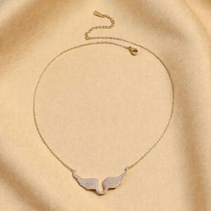 K&ouml;nnte beinhalten: Goldfarbene Halskette mit einem Anh&auml;nger in Form von Engelsfl&uuml;geln. Die Fl&uuml;gel sind mit kleinen, glitzernden Kristallen besetzt. Die Halskette wird vor einem hellbeigen Hintergrund pr&auml;sentiert.