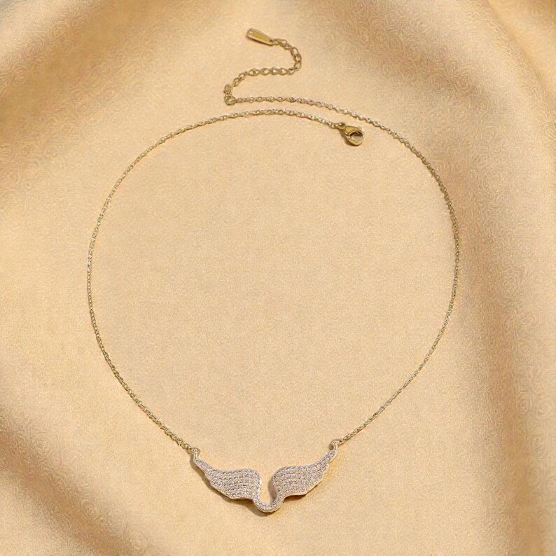 K&ouml;nnte beinhalten: Goldfarbene Halskette mit einem Anh&auml;nger in Form von Engelsfl&uuml;geln. Die Fl&uuml;gel sind mit kleinen, glitzernden Kristallen besetzt. Die Halskette wird vor einem hellbeigen Hintergrund pr&auml;sentiert.