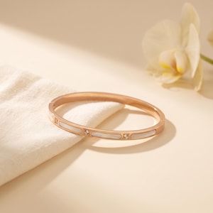 Op de afbeelding: Een scharnierende armband in roségoudkleur met afwisselende rechthoekige inlays van wit parelmoer en kleine, heldere, rondgeslepen fonkelende steentjes die in de metalen band met een gepolijste afwerking zijn gezet.