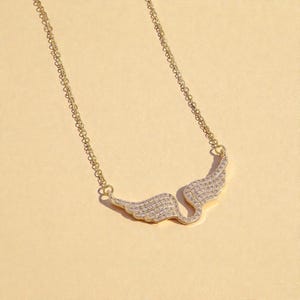 K&ouml;nnte beinhalten: Eine goldfarbene Halskette mit einem fl&uuml;gelf&ouml;rmigen Anh&auml;nger. Die Fl&uuml;gel sind mit kleinen, klaren Edelsteinen besetzt. Die Kette der Halskette ist filigran und ebenfalls goldfarben.