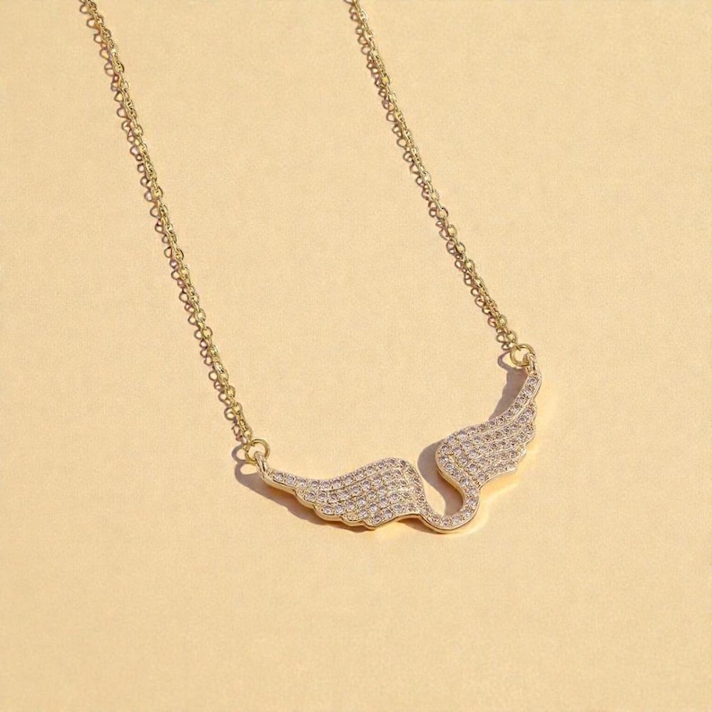 K&ouml;nnte beinhalten: Eine goldfarbene Halskette mit einem fl&uuml;gelf&ouml;rmigen Anh&auml;nger. Die Fl&uuml;gel sind mit kleinen, klaren Edelsteinen besetzt. Die Kette der Halskette ist filigran und ebenfalls goldfarben.