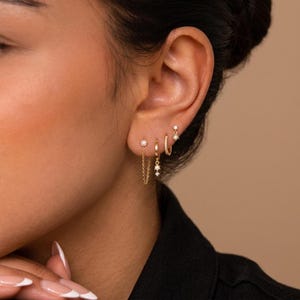 May include: A set of four gold earrings worn in a single ear. The set includes a small stud, a thin hoop, a hoop with a dangling star-shaped charm, and a stud connected to a delicate chain that loops back to the earlobe for a layered look.