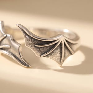 Pode incluir: Um anel aberto de tom prateado com dois designs sim&eacute;tricos de asas de morcego. O metal tem um acabamento polido nas bordas elevadas e uma apar&ecirc;ncia texturizada e escura nas &aacute;reas rebaixadas das estruturas das asas, criando detalhes.
