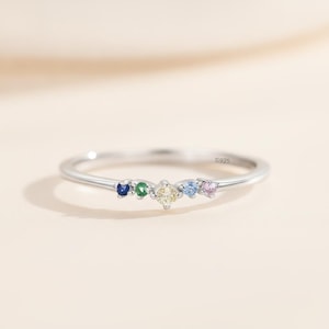 May include: A silver ring featuring a delicate band with an S925 hallmark. The centre of the ring is adorned with five small, round-cut gemstones in a row, displaying a vibrant colour gradient of dark blue, green, yellow, light blue, and pink.