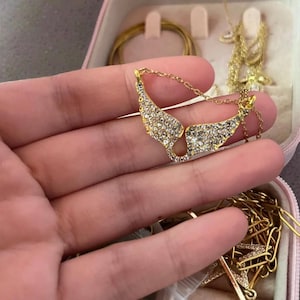 K&ouml;nnte beinhalten: Goldfarbene Halskette mit einem Anh&auml;nger in Form von Engelsfl&uuml;geln, besetzt mit glitzernden Kristallen. Die Halskette wird in einer Hand gehalten, im Hintergrund sind weitere goldene Schmuckst&uuml;cke zu sehen, was auf eine Schmucksammlung hindeutet.