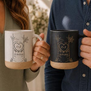 Peut inclure: Deux tasses en céramique, une blanc cassé et une noire, arborant chacune un motif de tronc d'arbre avec un cœur gravé contenant les lettres F+J et la date 03.26.2026. Les deux tasses ont une base beige texturée et une finition lisse.