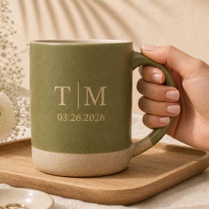 May include: A sage green ceramic mug with a speckled beige base, featuring the engraved letters T and M separated by a vertical line, with the date 03.26.2026 printed below in a serif font, held by a hand with a white sleeve.
