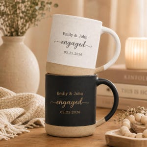 May include: Two ceramic mugs stacked vertically. The top mug is off-white with a speckled texture, featuring the text Emily & John engaged 03.25.2026. The bottom mug is matte black with the same text in gold, both featuring a tan unglazed base.
