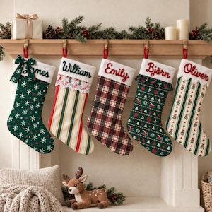 Op de afbeelding: Vijf gepersonaliseerde kerstsokken hangen aan een houten schoorsteenmantel. De sokken dragen de namen James, William, Emily, Björn en Oliver, met patronen van sneeuwvlokken, strepen, ruiten, sleeën en maretakken.