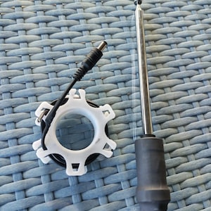 Può includere: Un'antenna argentata con base nera e cavo di alimentazione nero con un componente bianco a forma di ingranaggio. L'antenna è in posizione verticale. I componenti sono su una superficie blu intrecciata.