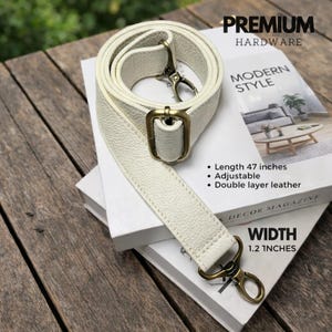 Puede incluir: Correa de bolso de cuero color crema con herrajes de latón envejecido. La correa mide 120 cm de largo y 3 cm de ancho, es ajustable y está hecha de cuero de doble capa. El texto "PREMIUM HARDWARE" y "MODERN STYLE" es visible.