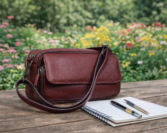 May include: A burgundy leather crossbody bag with a flap closure and adjustable strap sits on a wooden surface. A spiral-bound notebook and two pens are next to the bag. The background features a colorful flower garden.