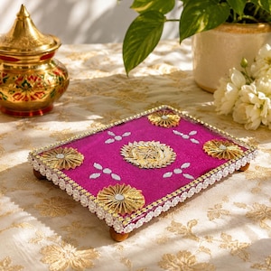 May include: A rectangular magenta tray featuring gold-toned circular embellishments at each corner and a central gold floral motif. The edges are decorated with white lace trim and small mirror accents are scattered across the surface. The tray stands on four small brown feet.