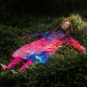 May include: A person wearing a colorful raincoat with pink leggings and sneakers lies in a bed of green foliage. The raincoat features a vibrant mix of pink, blue, and purple hues. The scene is set outdoors, with a dark background.
