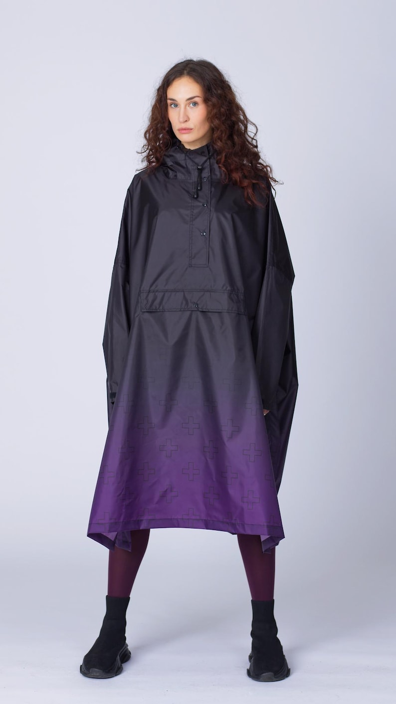 May include: Black hooded poncho with a purple ombre design. The poncho features a buttoned closure and a subtle cross pattern at the hem. The garment is worn with dark leggings and black ankle boots, creating a modern, stylish look.