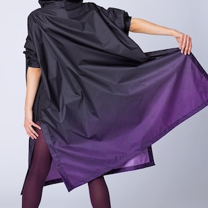 May include: Black hooded rain jacket with a purple ombre design and cross pattern. The jacket has short sleeves and a split hem. The model is wearing purple tights and black Steve Madden boots.