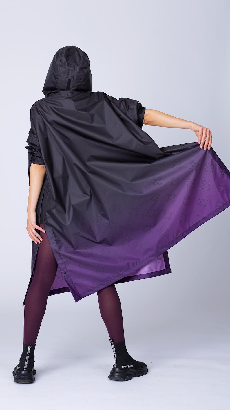 May include: Black hooded rain jacket with a purple ombre design and cross pattern. The jacket has short sleeves and a split hem. The model is wearing purple tights and black Steve Madden boots.
