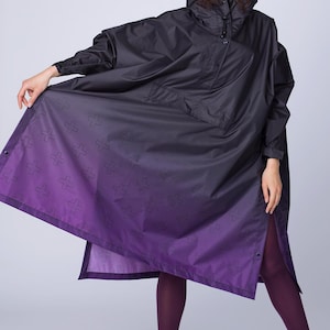 May include: Black hooded poncho with a purple ombre design. The poncho features a button closure and a subtle cross pattern on the lower portion. The model is wearing black boots and purple leggings. The poncho is a stylish outerwear option.