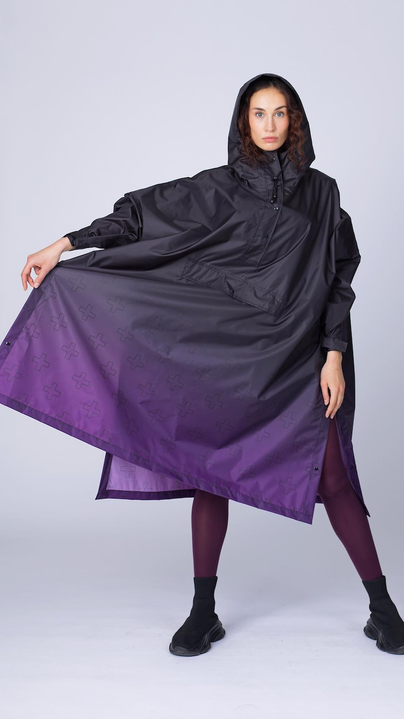 May include: Black hooded poncho with a purple ombre design. The poncho features a button closure and a subtle cross pattern on the lower portion. The model is wearing black boots and purple leggings. The poncho is a stylish outerwear option.