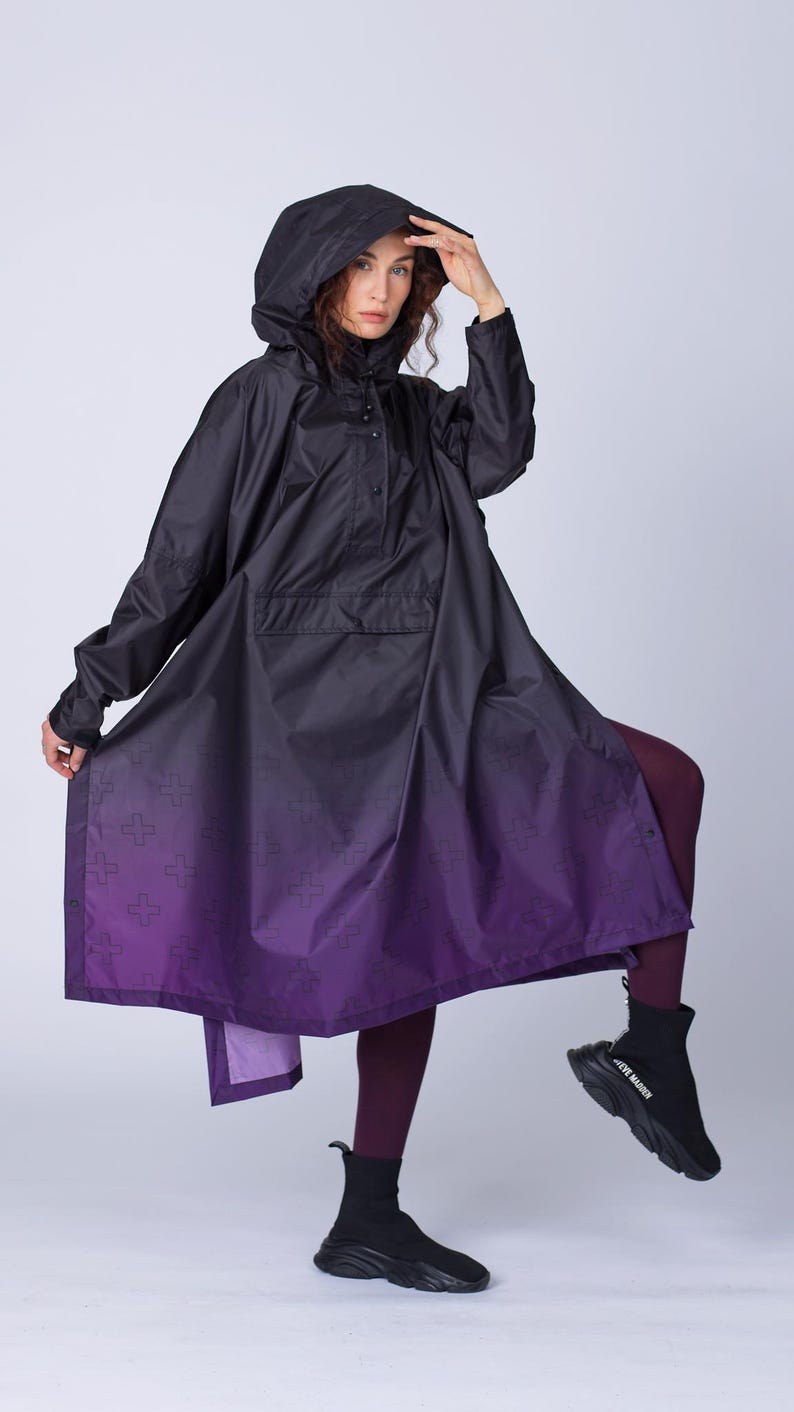 May include: A black hooded poncho-style raincoat with a purple ombre effect at the hem. The raincoat features a front pocket and a cross pattern on the purple section. The model is wearing black ankle boots.